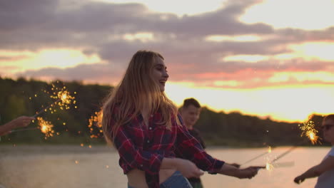 The-blonde-girl-in-red-plaid-shirt-and-denim-shorts-is-dancing-with-big-bengal-lights-in-her-hands-on-the-beach-with-her-friends.-This-is-beautiful-summer-evening-on-the-lake-coast-near-the-forest.